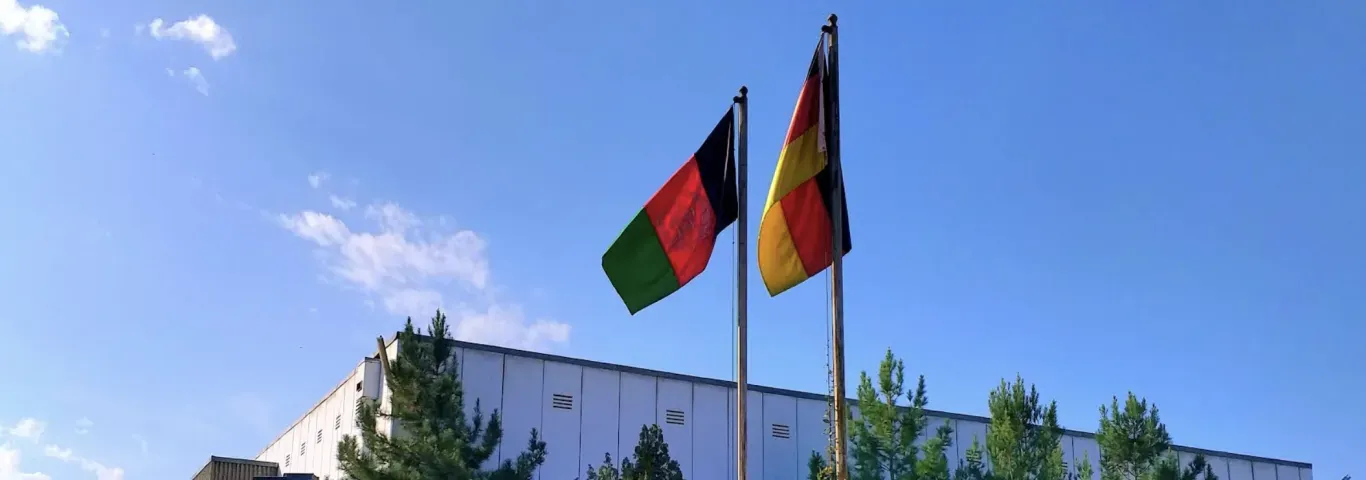 This photograph captures the flags of Afghanistan and Germany waving together against a clear blue sky above a building.