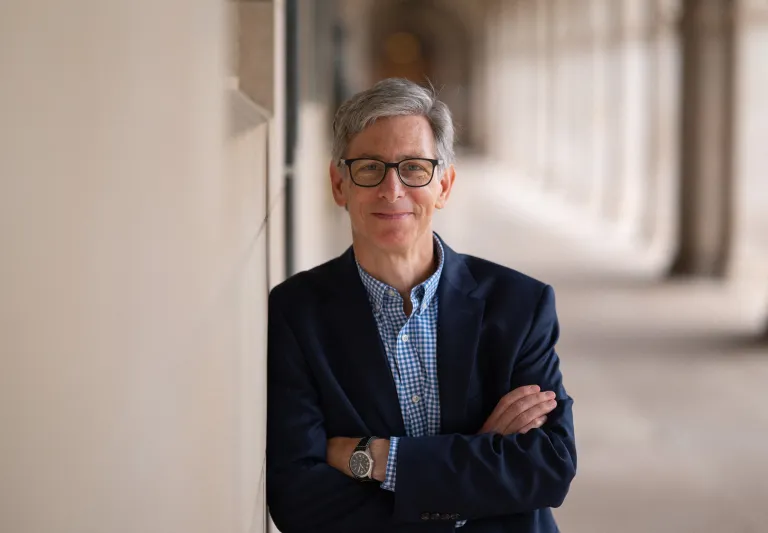 Peter Kastor leans against a wall and smiles at the camera. He is wearing dark-brimmed glasses and a suit.
