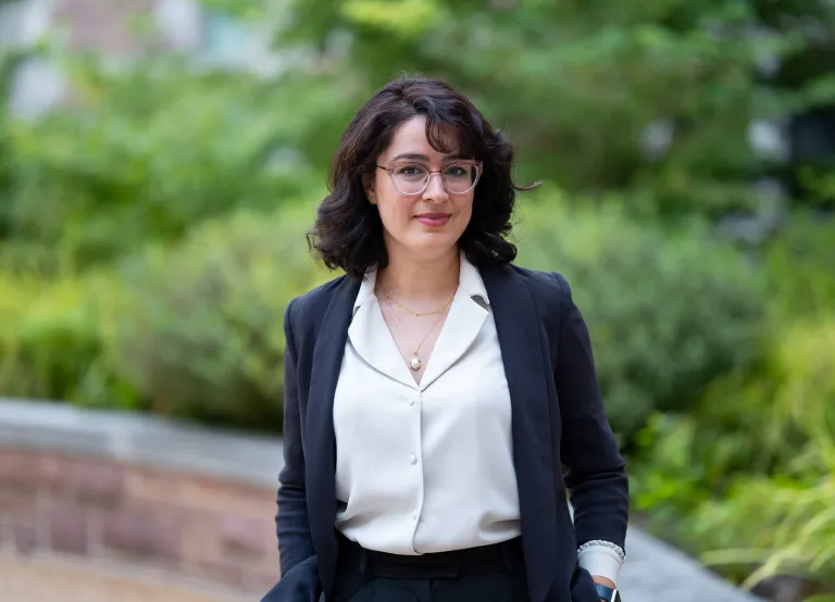 Image of Marjan Wardaki - she is wearing a white button-up shirt with a black long-sleeve blazer. She has wide-brimmed glasses. Wardaki smiles at the camera.