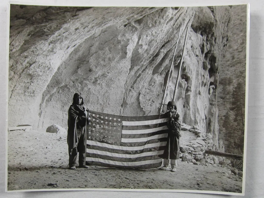 "Two Pueblo individuals hold an American flag, an image from the early 1900s."